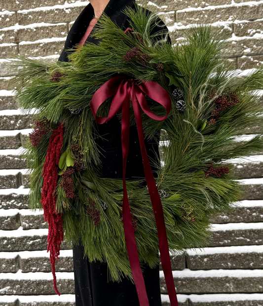 Person holding a green wreath with red ribbon against a snowy brick wall.