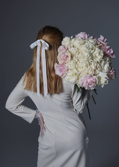 A person from behind holding a large bouquet of light pink and white roses with a ribbon tied in a bow on the head.