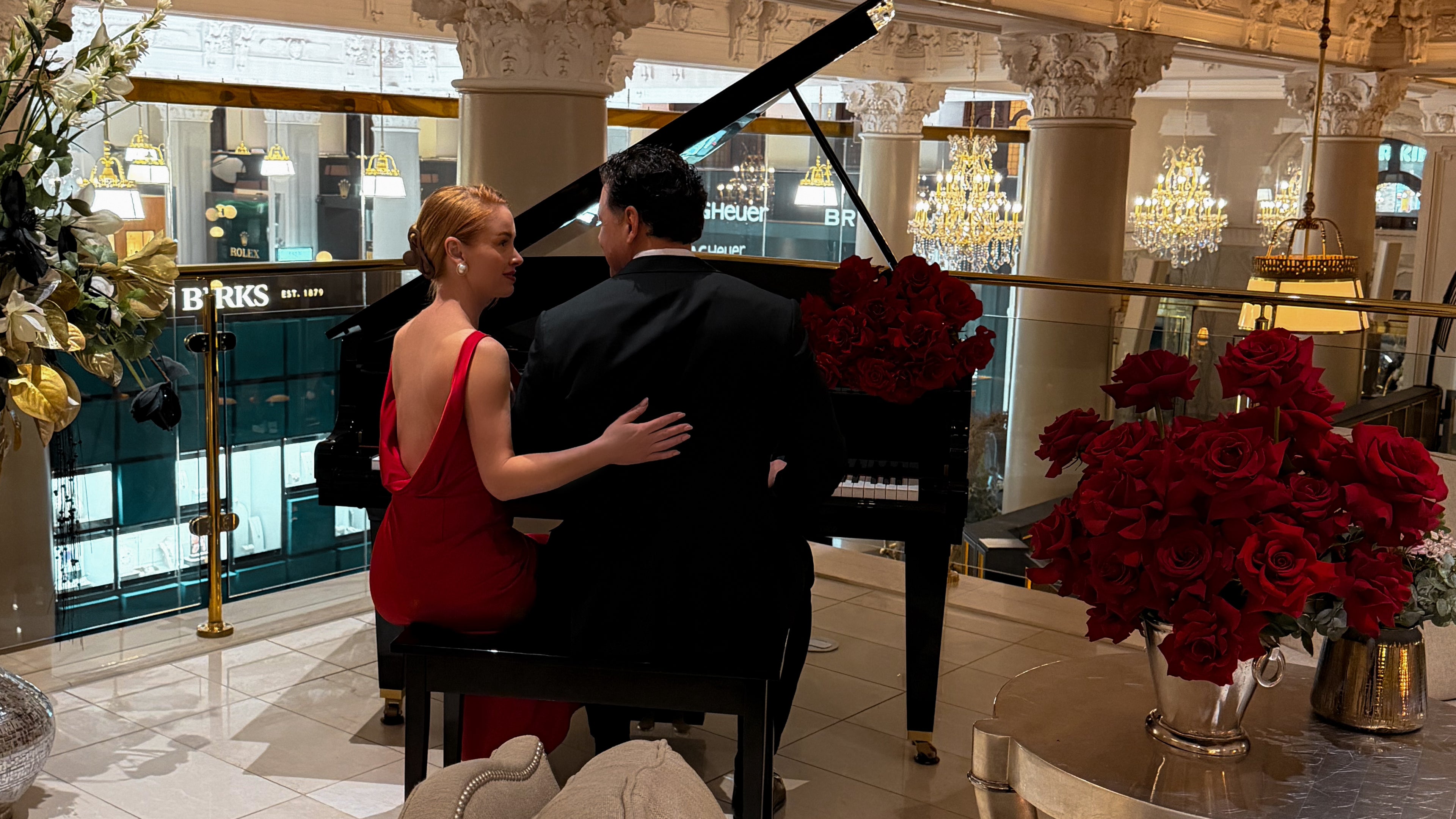 Man and woman by a grand piano in an elegant indoor setting with flowers and a blurred background.